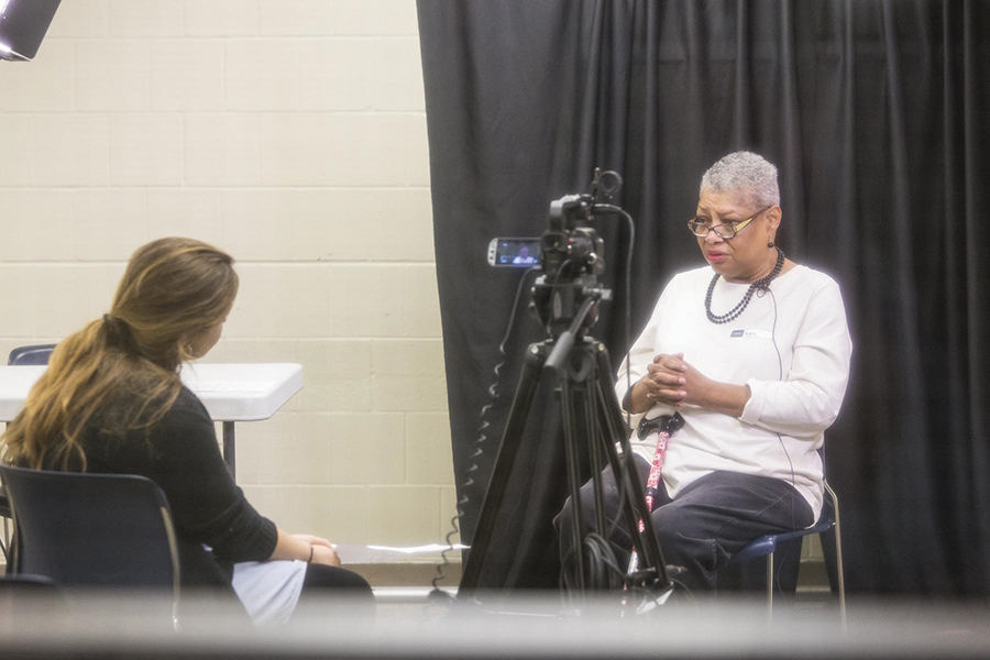 This picture is taken through a window. Williams is sitting in front of a black curtain. A student sits behind a camera recording her interview.