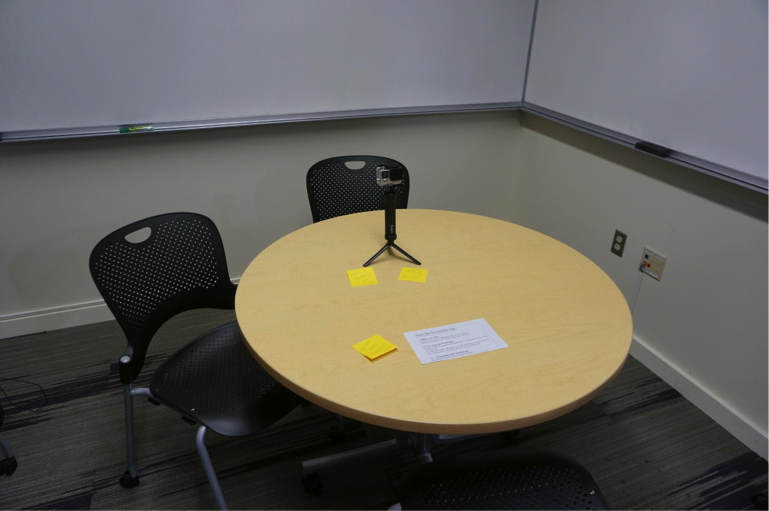 Figure 1: An image of a small room with two empty chairs and a table. On the table is a video camera on a tripod, and a list of questions that contain the prompts for the video. 