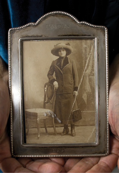 The hands of Suzanne Pred Bass hold an antique framed black-and-white image of her great aunt Rosie Weiner, an older white woman wearing a dark grey suit with a black blouse and a wide brim grey hat from the 1930s.