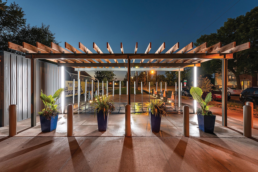 Nighttime view of the Rondo Plaza. The lights illuminate the art installation underneath a wooden pergola. Four large, blue planters are placed equal distance apart in front of the pergola.