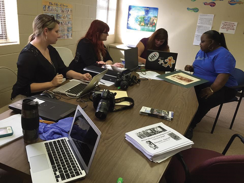 Three students are gathered around a conference table with Youngblood. They are sitting with their laptops, having and conversation and working.