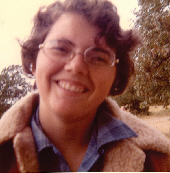 Close up and somewhat blurry photo of Jeanne Barrett, wearing glasses and a shearling jacket, with a tree in the background.