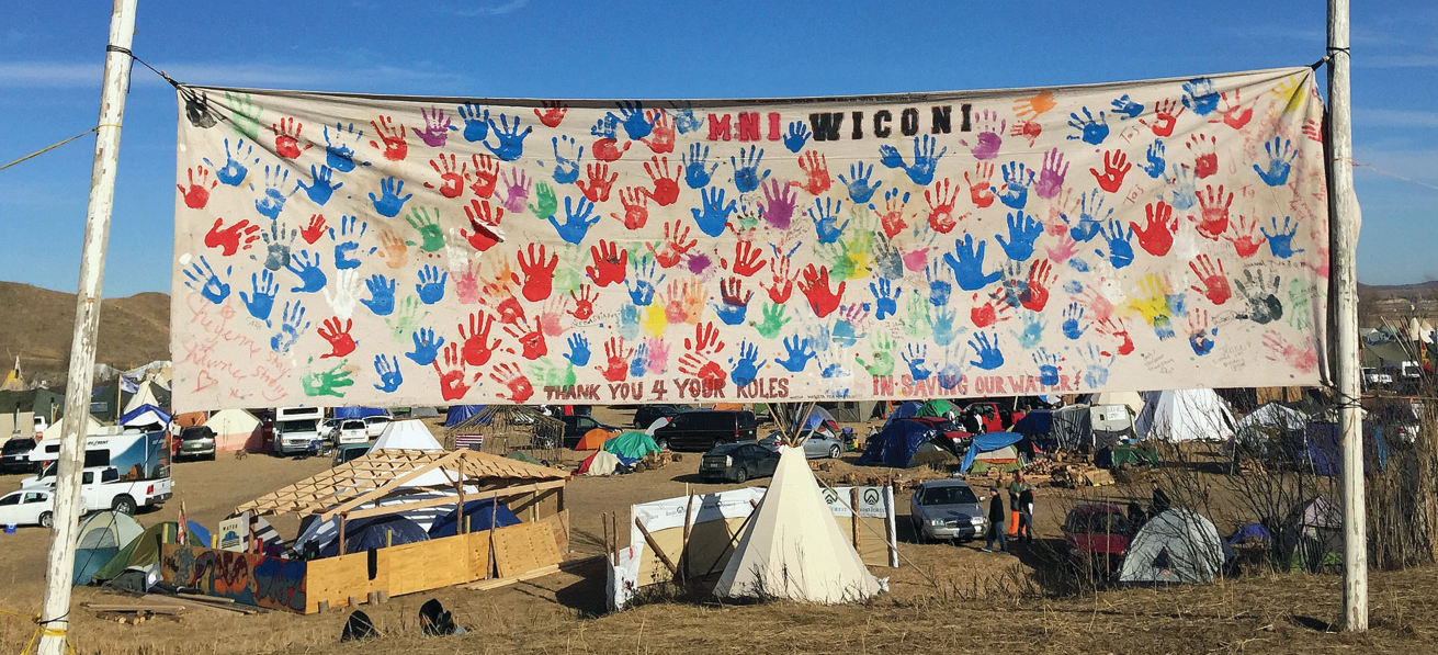 A camp settlement shows several vehicles, tents, and a teepee. A large banner covered with brightly colored handprints reads “MNI WICONI” and “Thank You 4 Your Roles in Saving Our Water”.