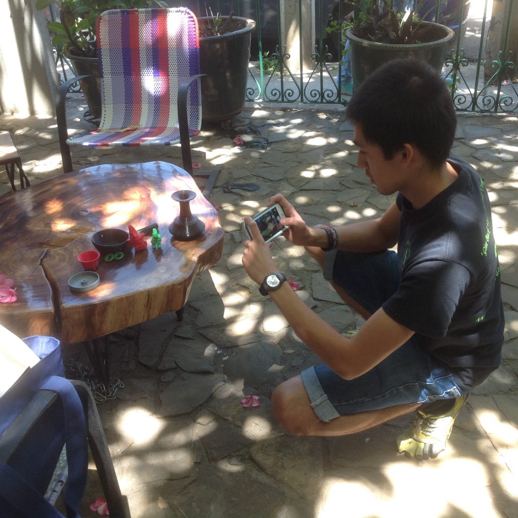 Fig. 11 – Young man with a camera kneels by a table with several plastic objects on it, taking a photograph of them. 