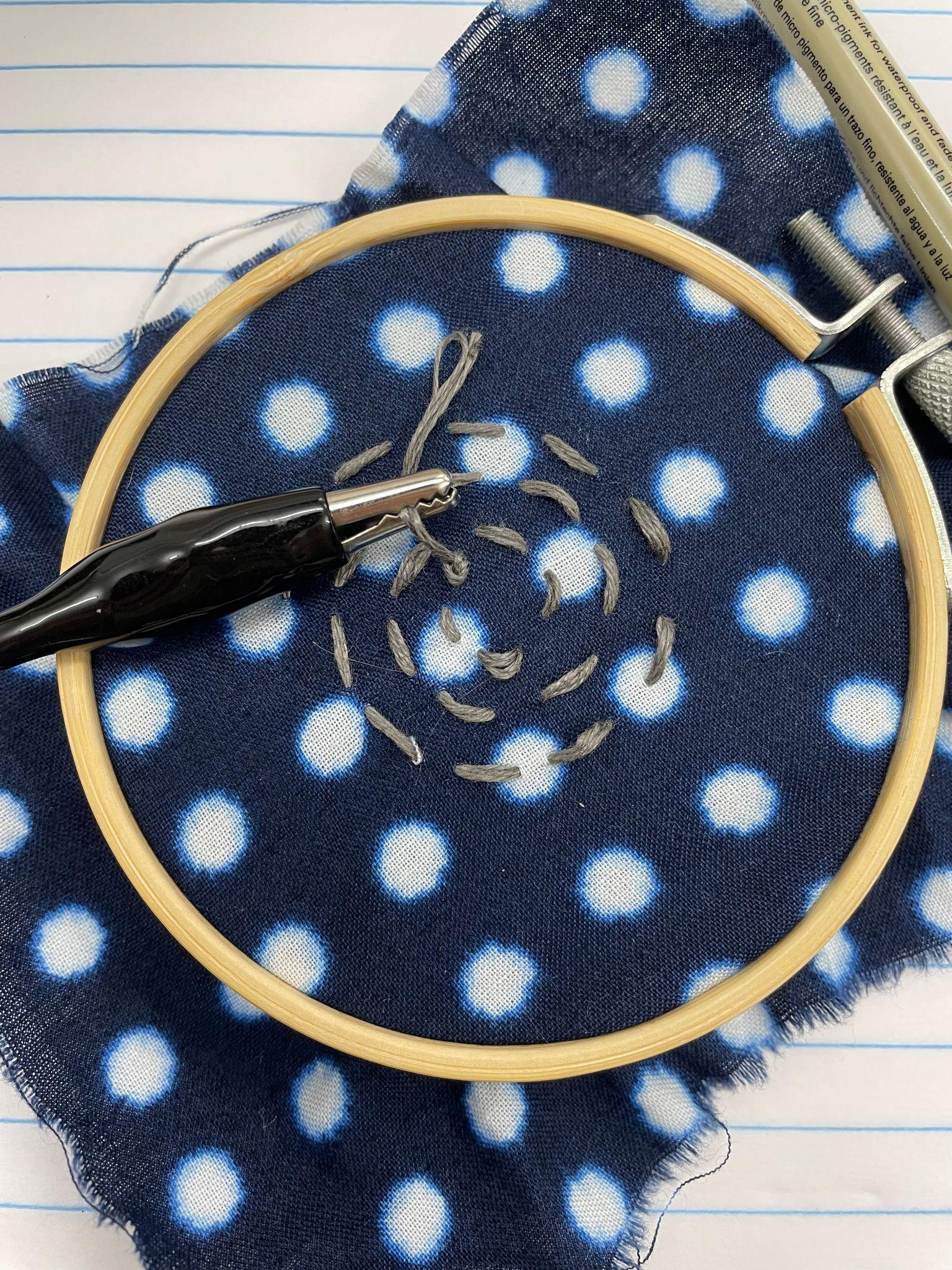 An embroidery hoop with blue and white fabric in it. Gray steel thread has been stitched in a spiral on the fabric, and the end of the thread is connected to an alligator clip.
