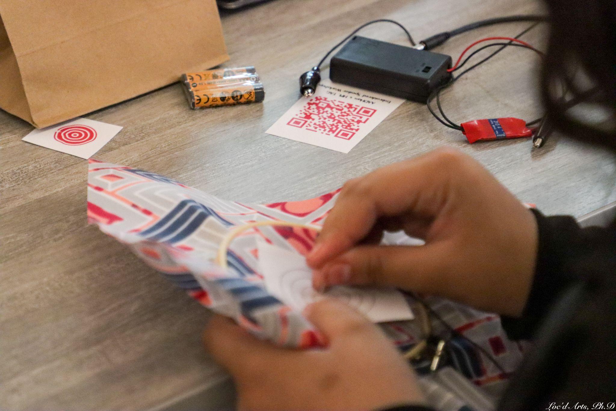A student’s hands holding an embroidery hoop with gray, blue, and red fabric. A battery pack, alligator clips, batteries, and a piece of paper with a red QR code are visible on the table in the background.