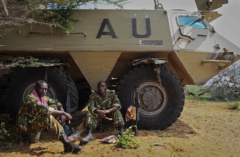 Color photo of men in camouflage fatigues sitting in front of a tank that has the letters AU stenciled on it.  