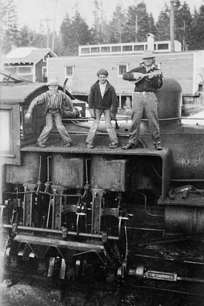 Three male laborers of various ages are posed on top of a steam locomotive, striking a variety of poses for the camera. The man on the right appears to be holding a small guitar, and the most left person appears to be a younger boy.