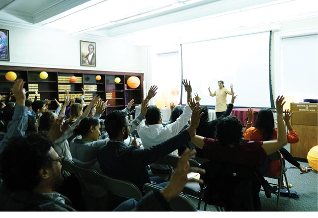 This photo shows a South Asian woman facilitating a workshop with several South Asian participants. The participants, or the audience, mirror the Facilitator’s gesture, leading everyone in the bright room to have two hands raised.