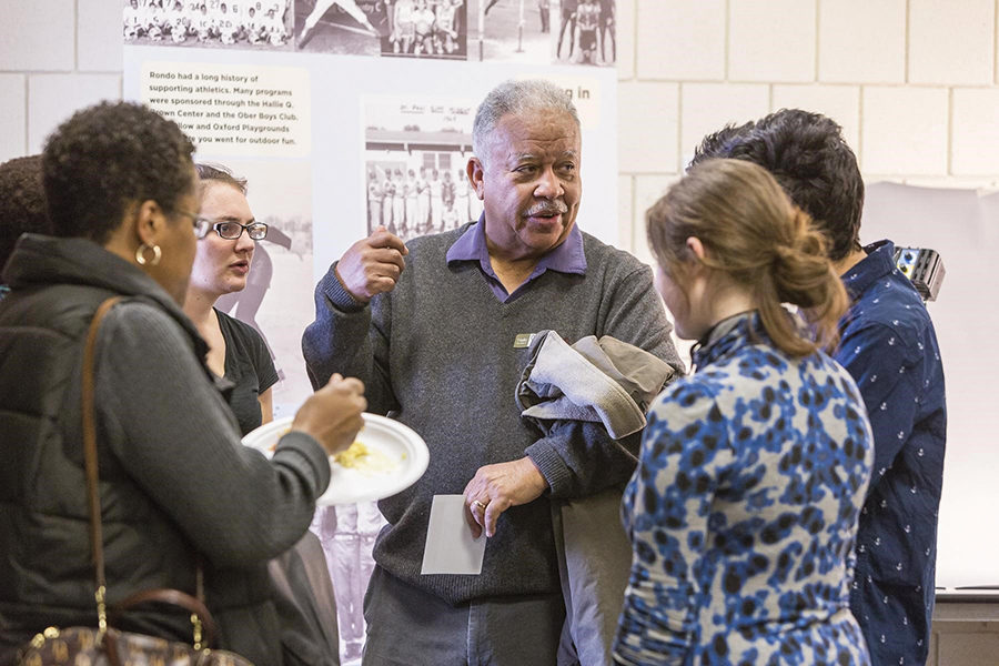 Six people are gathered in a circle in front of a placard telling Rondo’s history. Three of the people are students in the History Harvest class; three are Rondo residents. One of the participants is eating lunch.