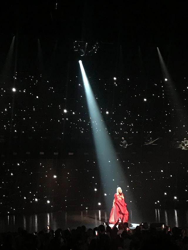A performer in a dramatic red gown sings on stage, illuminated by a single spotlight. The background is dark with scattered star-like lights.
