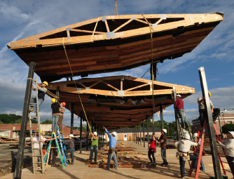 Pavilion is wood. One piece is being hoisted into place by a crew of workers.