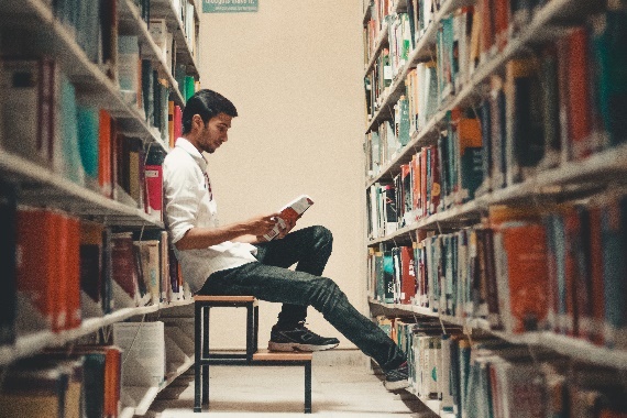 Photograph: person reading and sitting on a stool between shelves of books.
