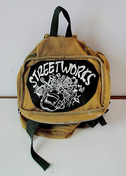 The brown backpack has a black and white patch that says STREETWORKS on it with line drawings below of various items like toothbrushes, bus passes, matches, thread, needles, and socks.