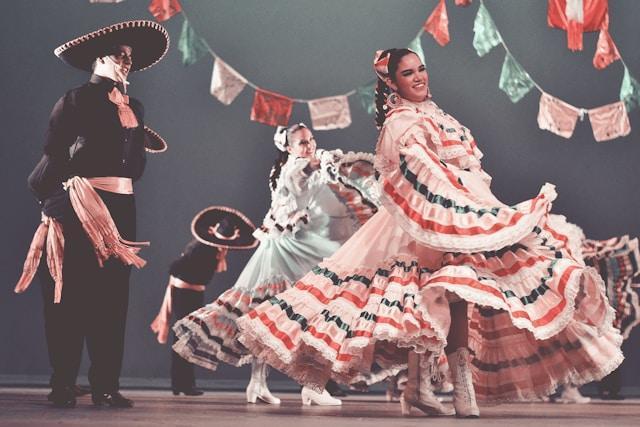Dancers perform in traditional Mexican attire, with women in colorful dresses and men in sombreros. They are surrounded by festive hanging flags, creating a vibrant, cultural atmosphere.