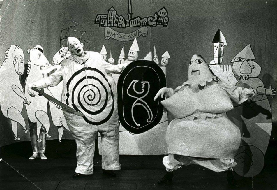 A group of people in costume on stage acting out a surreaslist play. One actor holds a sword and shield. 

