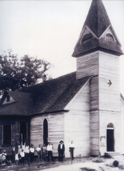 This historic photograph depicts a dozen or more congregants standing to the left of the church. The church is wooden with a single, large room and a tall bell tower in the front.