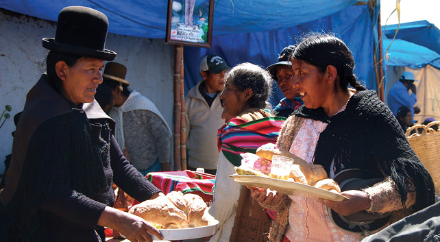 Pictured here is a market scene where an indigenous woman in La Paz, Bolivia purchases fresh bread from an indigenous Bolivian man. Other indigenous people are also shopping in the background.