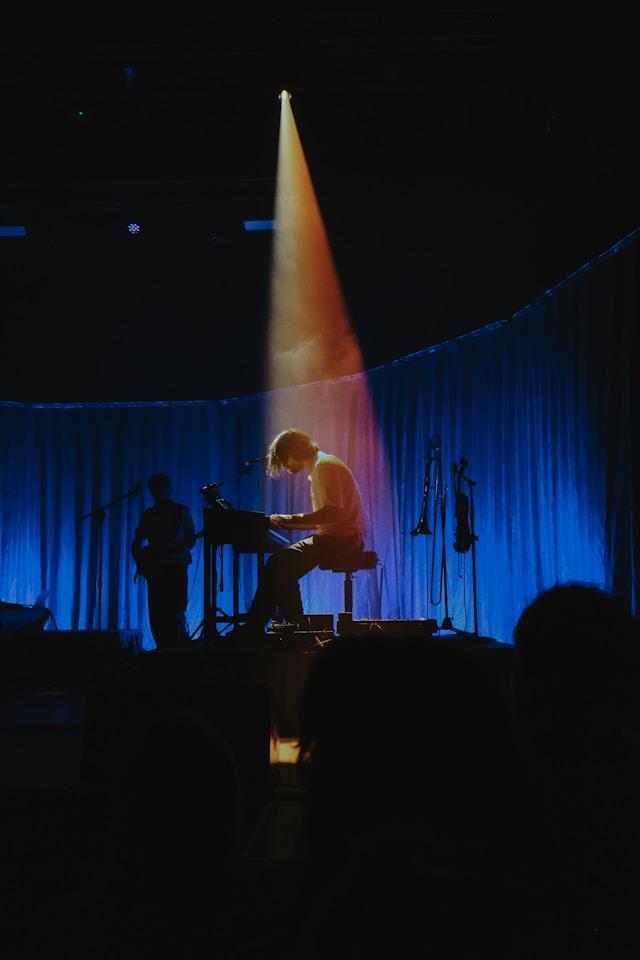 A musician sits at a keyboard on a dimly lit stage. A spotlight shines on them, creating a dramatic ambiance. Blue curtains and silhouettes of other instruments and stands are visible in the background.