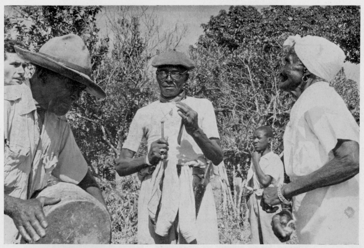 Photo of percussionsist, including a man playing a drum and another playing a metal hoe blade with an iron nail, along with a woman dancing, and other individuals in the background.