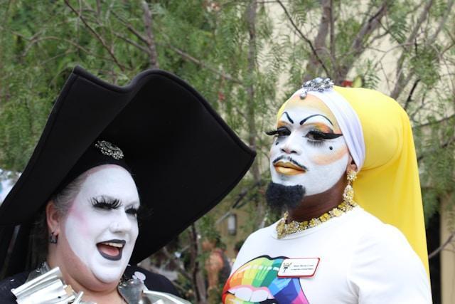 Two people with elaborate makeup and costumes pose outdoors. One wears a large black hat and white face paint, while the other has a yellow headdress and a colorful shirt. They stand in front of trees, smiling. These people are a part of the Sisters of Perpetual Indulgence.