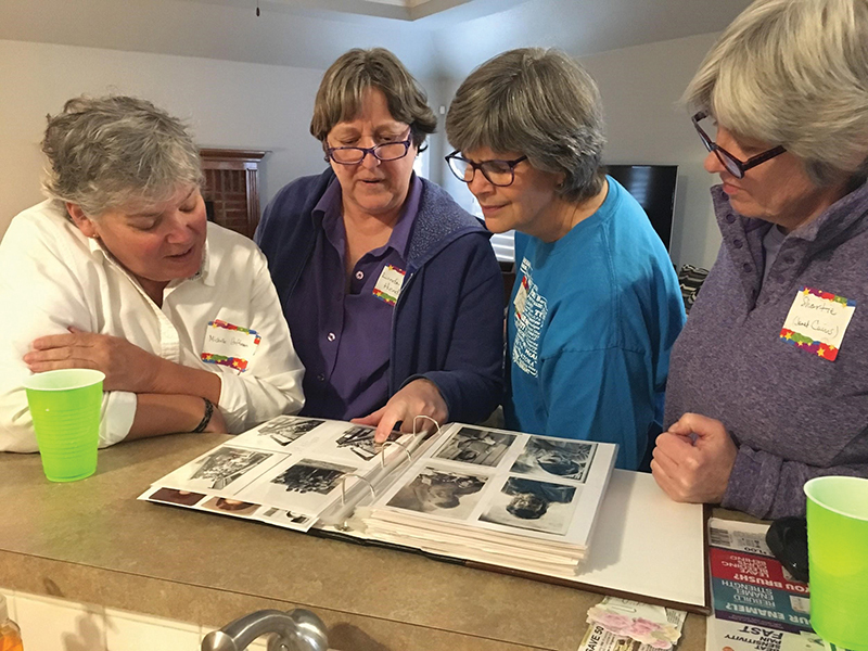 Four middle-aged women are standing at a kitchen counter looking at a Girl Scout photo album together.