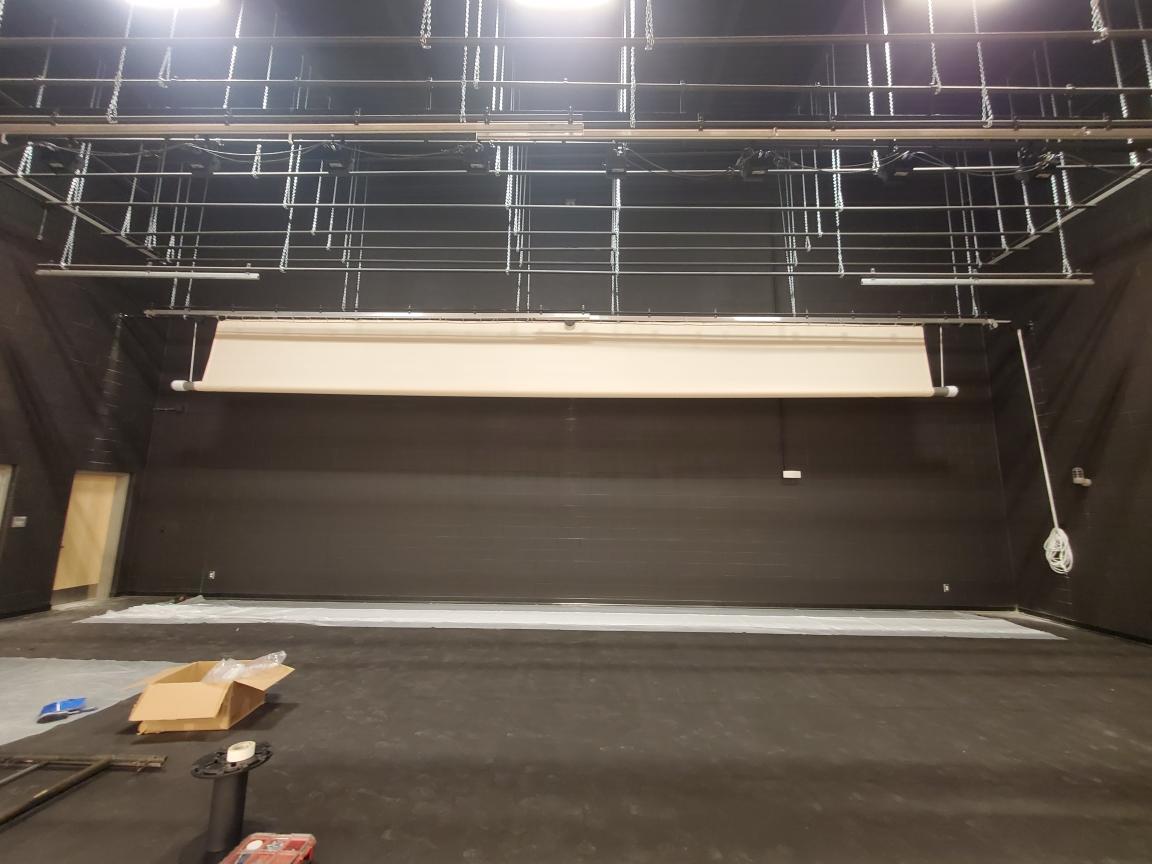 A group of people sitting around a campfire in a forest at night. The fire casts a warm glow on their faces. Background includes trees and hints of tents, suggesting camping.