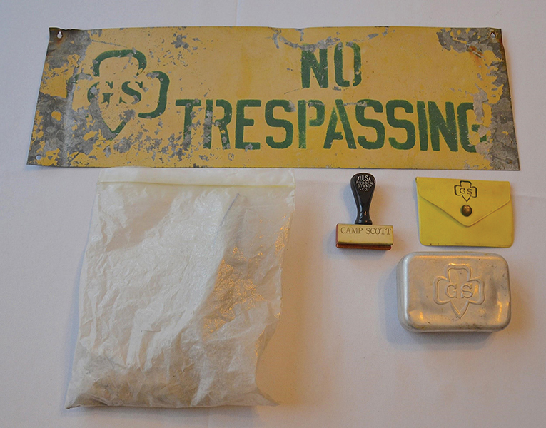 Green and yellow metal sign that reads “No Trespassing” with the Girl Scout Trefoil logo; a plastic baggie that contains ashes from a campfire in the 1970s; a Camp Scott rubber stamp; a plastic yellow and green Girl Scout coin purse with a brass snap; a tin soap container with the Girl Scout Trefoil embossed on it.