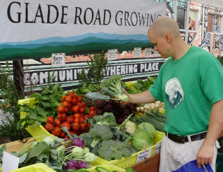 A man in green is holding Broccoli; standing in front of a produce stand that reads “Glade Road Growing.”