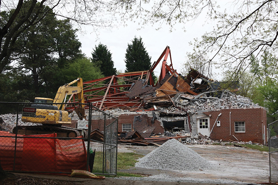 Color photograph of the demolition of the old brick church building. The picture is taken from a distance behind a demolition fence.
