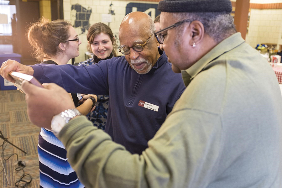 Two men stand in a community center room holding a large picture frame. Two students are behind them chatting.