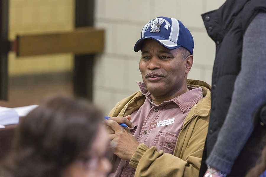 Melvin Carter, Jr. smiles during a conversation. He’s wearing a blue hat, a pink button-down shirt, and a tan jacket. A couple other figures surround him but are out of focus.
