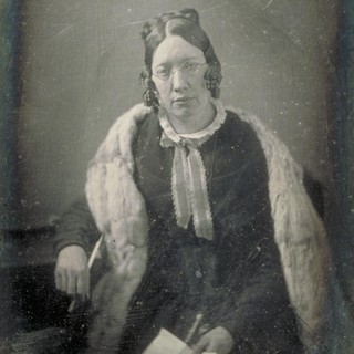 A faded and slightly cracked photo portrait of Catherine Beecher. She has a light-colored scarf across her shoulders, a dark dress, and a light-colored smaller neck-scarf. She is holding a piece of paper in her lap in one hand, and the other arm is rested on a table while holding a pen. The front/top of her hair has two neat space buns on either side of her head and the rest of her hair coils along her cheeks in a sort of short bob. She is looking directly at the camera with a very bored or exhausted facial expression.  Photo Credit: possibly by W & F Langenheim (1848). Source: Wikimedia Commons.