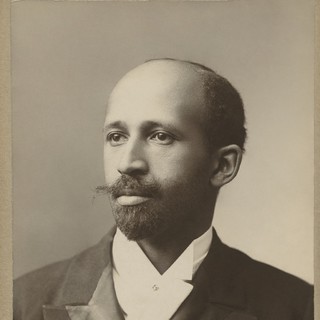 A sepia-toned photographed portrait of W.E.B DuBois. He is wearing an old-fashioned looking suit. He has light-brown skin, an English-style moustache, and a balding head. He is looking off to the left. Photo Credit: E. Purdy, 1907 (National Portrait Gallery)