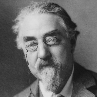 A gray scale photo portrait of Sidney Webb in a suit. He is wearing small oval-shaped classes with a chain attached on the left side.