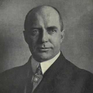 A grayscale photo portrait of W.I. Thomas. He is wearing a dark suit with a tie. His thinning hair is neatly trimmed around his head. He is focusing intently toward the right side of the photographer. Photo Credit: Photographer Unknown. Date: no later than 1907. Source: Wikimedia Commons. 