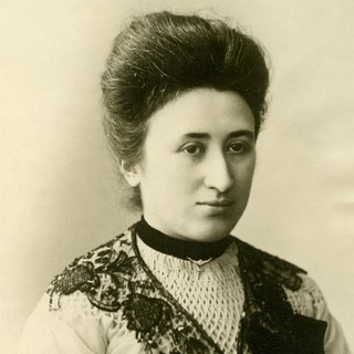 A sepia-toned photograph portrait of Rosa Luxemburg. She is wearing a high-neck dress with a v-neck-shaped lace detail as an accent. Her hair is neatly and voluminously coifed into an up-do. She is looking slightly to the right of the camera and she looks a bit bored or disinterested.  Photographer: Karl Pinkau. Year: 1900. Source: Wikimedia Commons