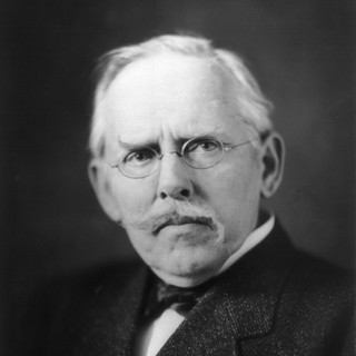 A grayscale photograph portrait of Jacob Riis. He is wearing a suit and a bowtie. He has round glasses and a moustache that flares upwards on the left side and downwards on the right side. His hair is gray and combed over neatly. He is looking sternly and seriously at the camera. Photo Credit: Pirie MacDonald (1906) Photo Source: Wikimedia Commons