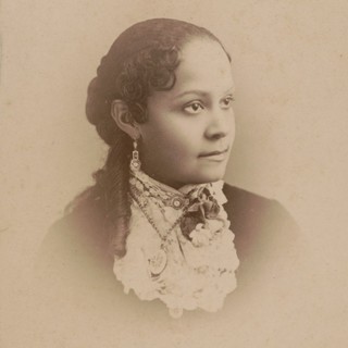 A sepia-toned photographed portrait of Fannie Barrier Williams. She is pensively looking to the right. She has neatly curled hair that rests on her shoulder and frames her face in bangs. She is wearing large and ornate earrings. Her shirt collar is frilly and covers her neck. Photo Credit: Paul Tralles, 1885. Source: commons.wikimedia.org